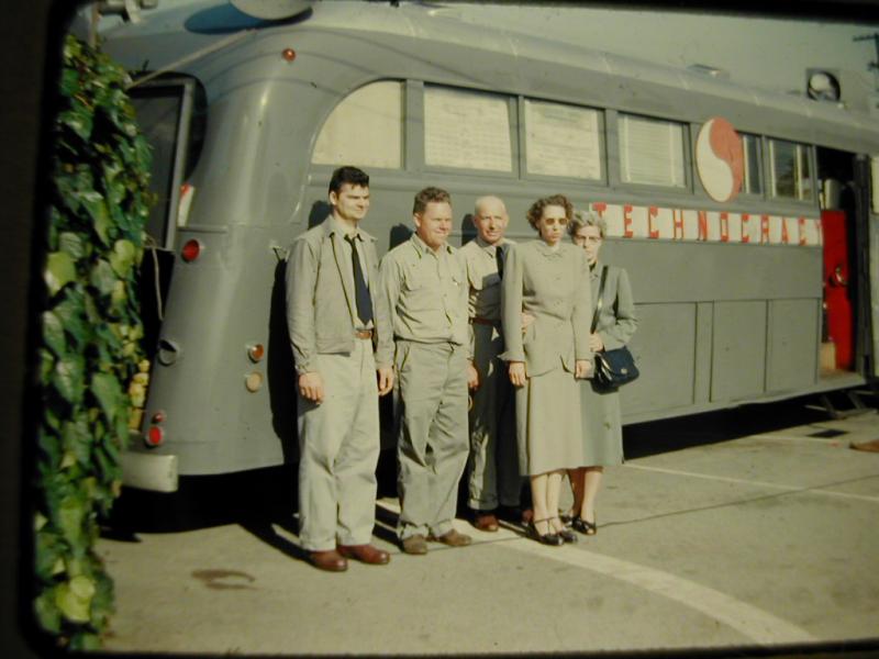 Three men in grey uniforms and two women in grey suits stand in front of a grey Technocracy sound car bearing the red and white Monad.